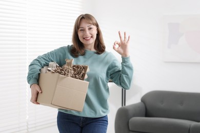 Photo of Recycling. Smiling woman holding cardboard box with different waste paper and showing OK gesture indoors