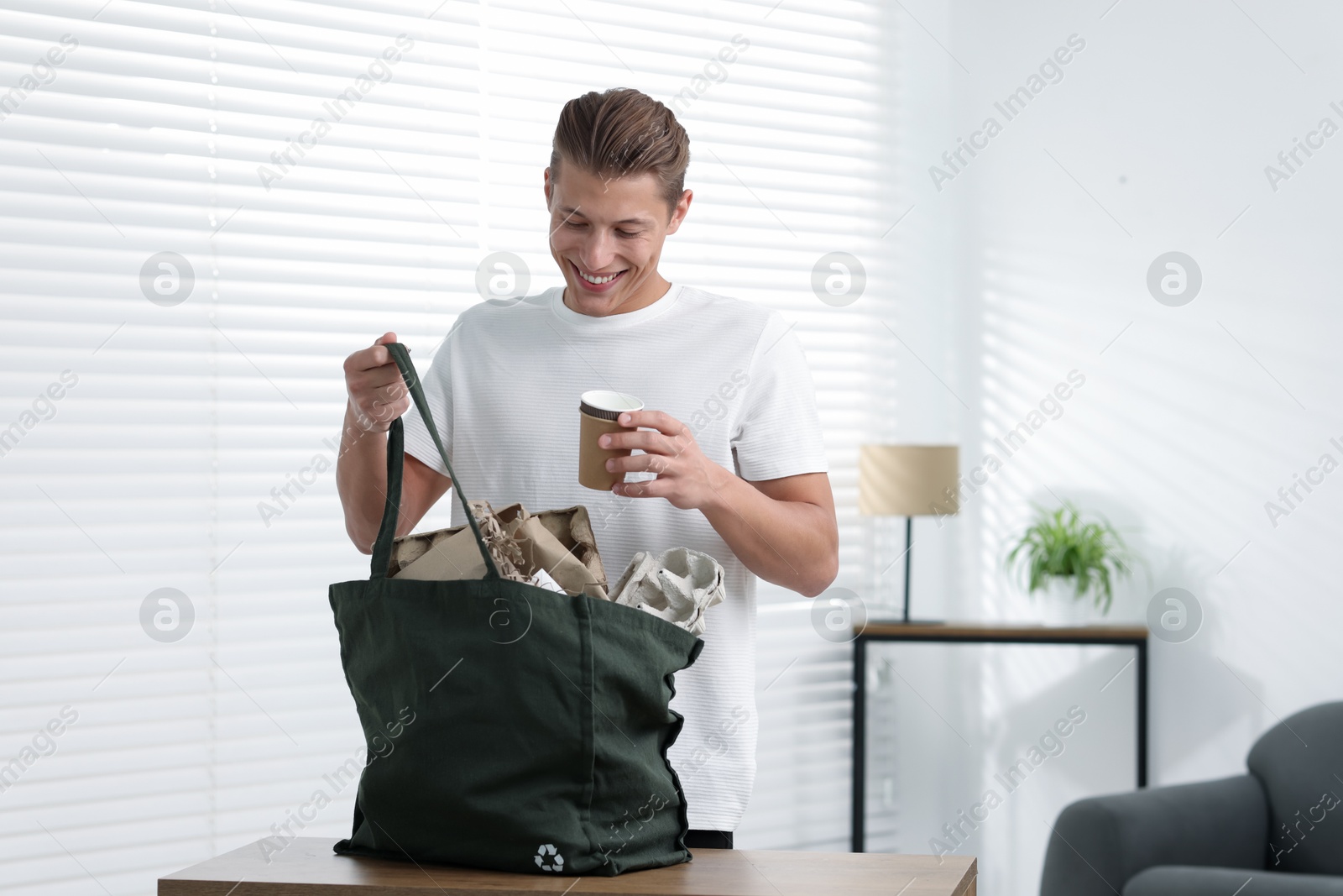 Photo of Recycling. Smiling man putting waste paper into bag at wooden table indoors