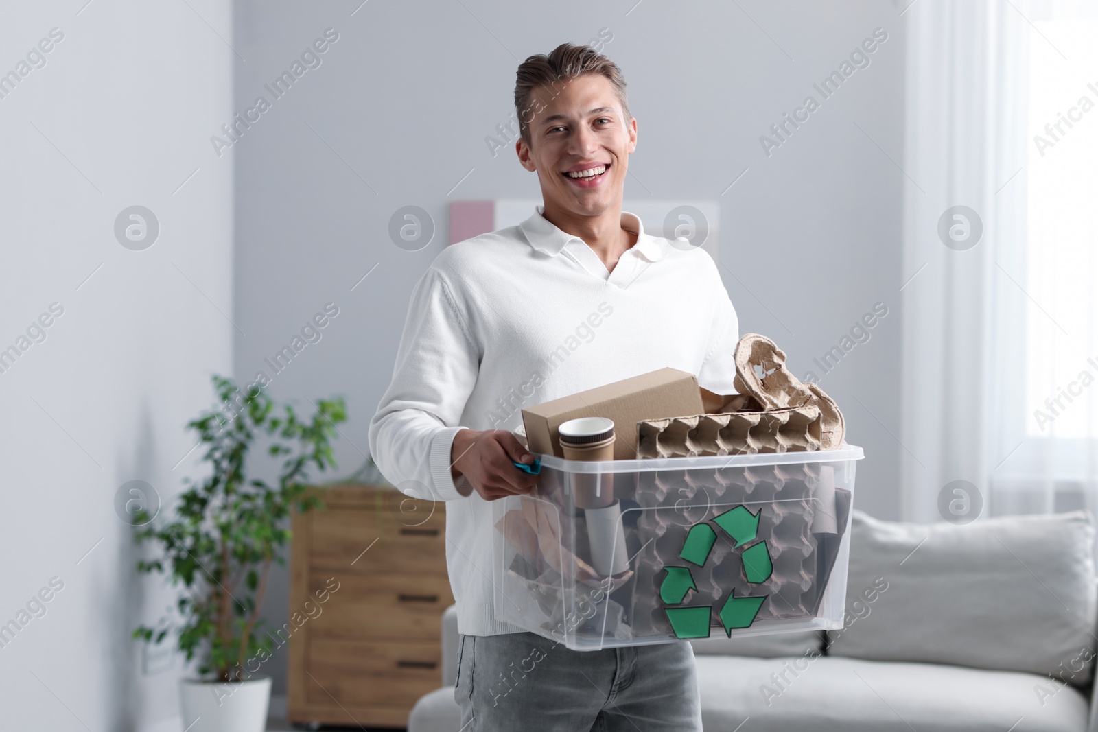 Photo of Recycling. Smiling man holding plastic box with different waste paper indoors