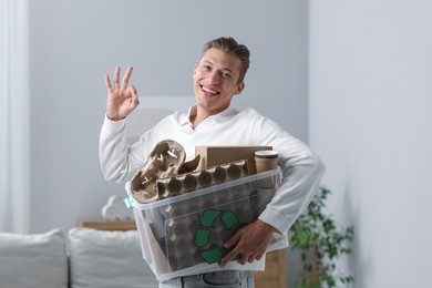 Photo of Recycling. Smiling man holding plastic box with different waste paper and showing OK gesture indoors