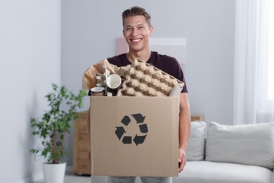 Photo of Recycling. Smiling man holding cardboard box with different waste paper indoors