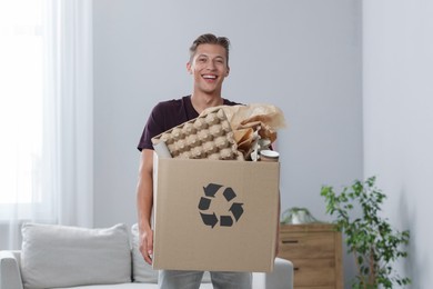 Photo of Recycling. Smiling man holding cardboard box with different waste paper indoors