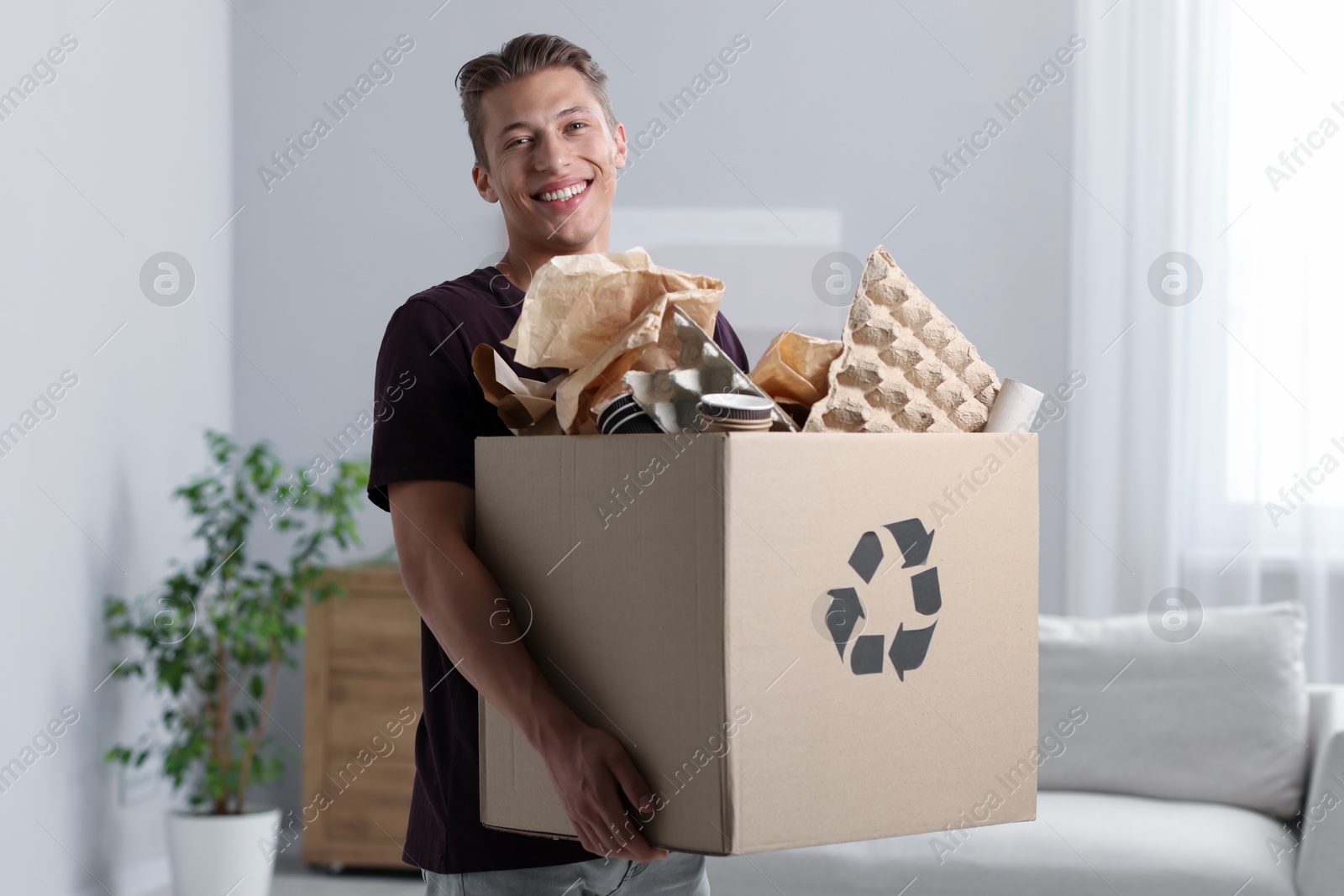 Photo of Recycling. Smiling man holding cardboard box with different waste paper indoors