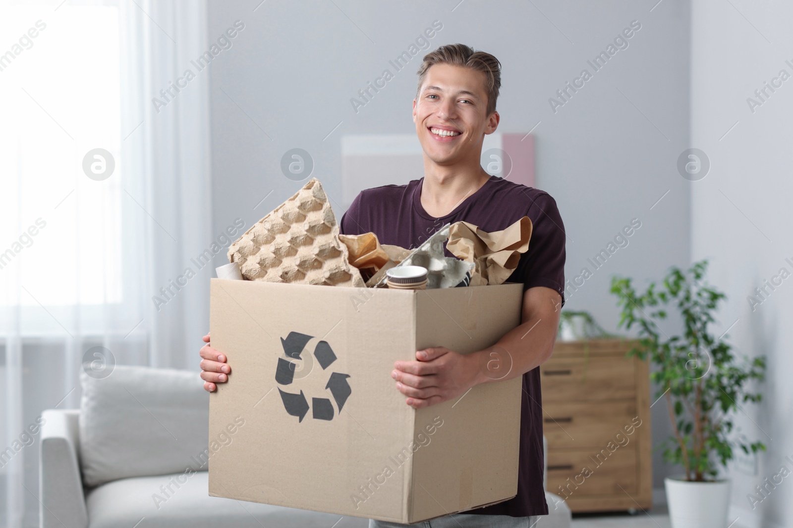 Photo of Recycling. Smiling man holding cardboard box with different waste paper indoors