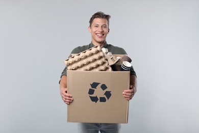 Photo of Recycling. Smiling man holding cardboard box with different waste paper on light background