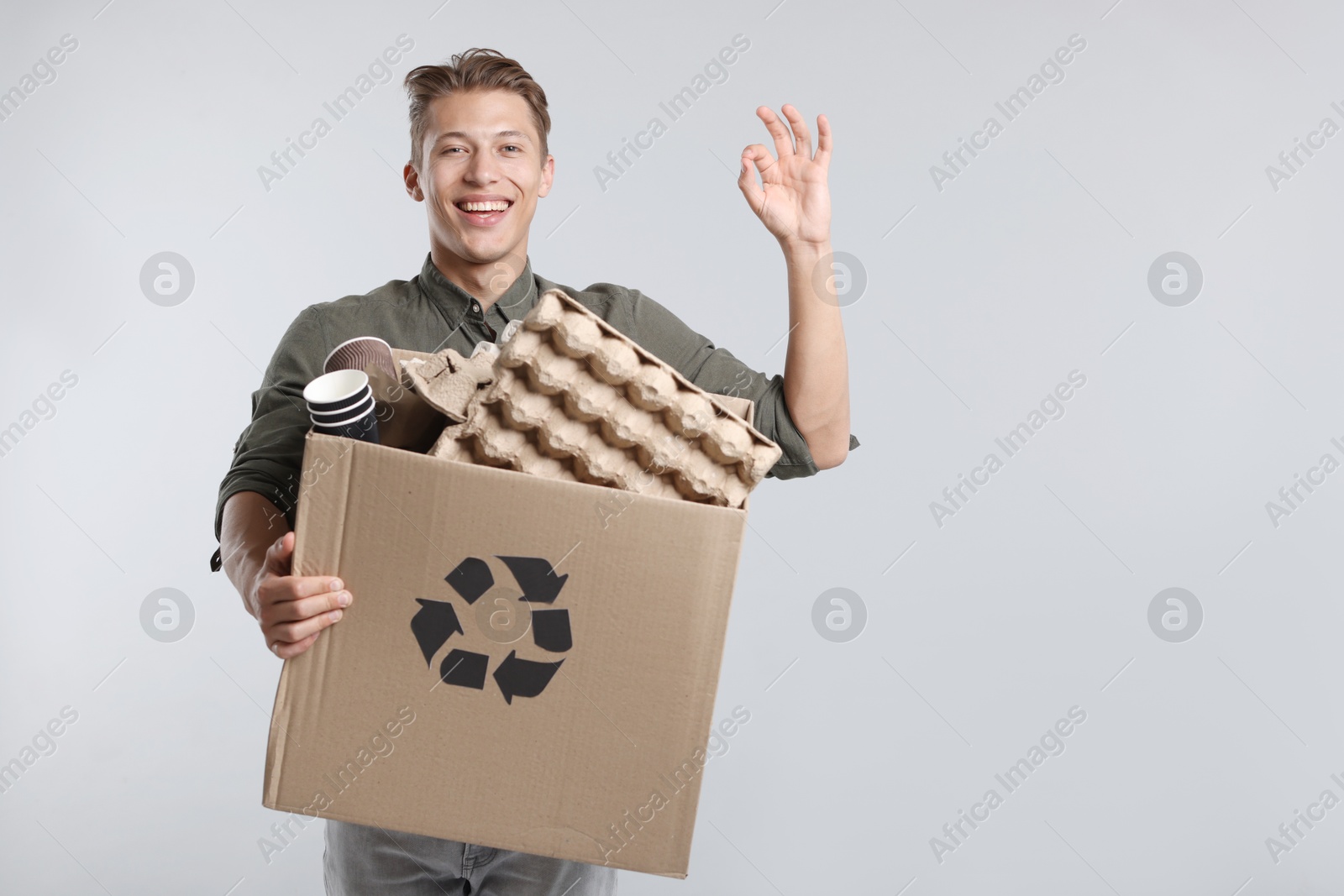Photo of Recycling. Smiling man holding cardboard box with different waste paper and showing OK gesture on light background. Space for text