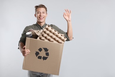 Photo of Recycling. Smiling man holding cardboard box with different waste paper and showing OK gesture on light background. Space for text