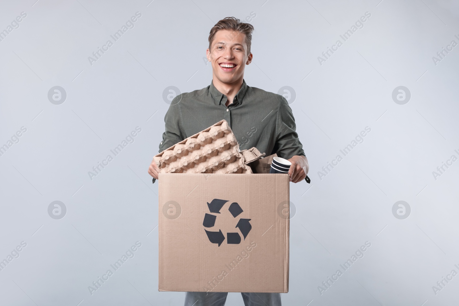 Photo of Recycling. Smiling man holding cardboard box with different waste paper on light background