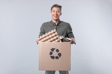 Photo of Recycling. Smiling man holding cardboard box with different waste paper on light background