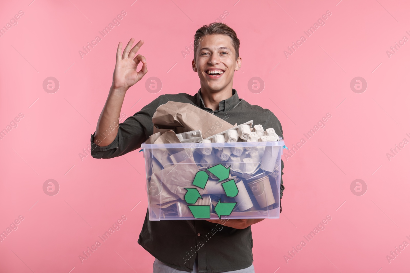 Recycling. Smiling man holding plastic box with different waste paper and showing OK gesture on pink background Photo of Recycling. Smiling man holding plastic box with different waste paper and showing OK gesture on pink background