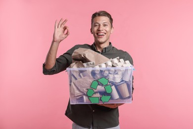Photo of Recycling. Smiling man holding plastic box with different waste paper and showing OK gesture on pink background