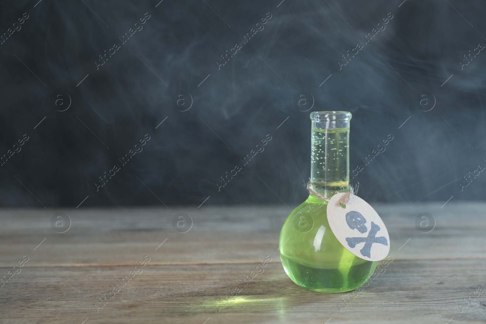 Photo of Flask of poison with warning sign on wooden table against dark background with smoke, space for text