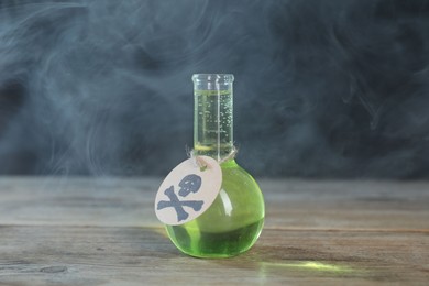 Photo of Flask of poison with warning sign on wooden table against dark background with smoke