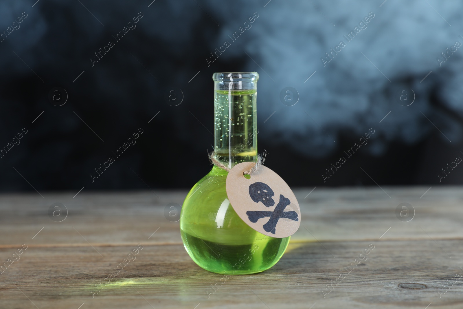 Photo of Flask of poison with warning sign on wooden table against dark background with smoke