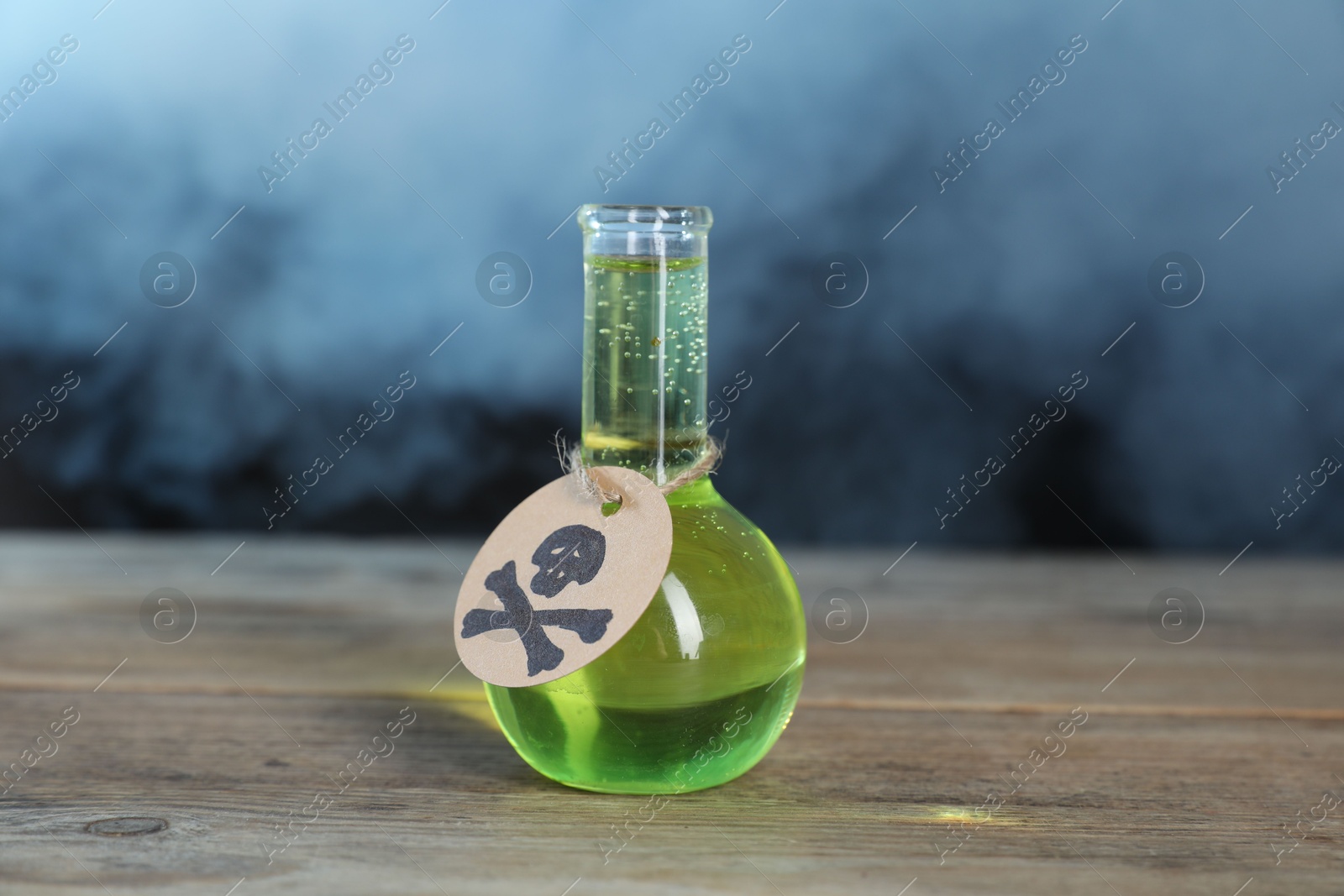 Photo of Flask of poison with warning sign on wooden table against dark background with smoke