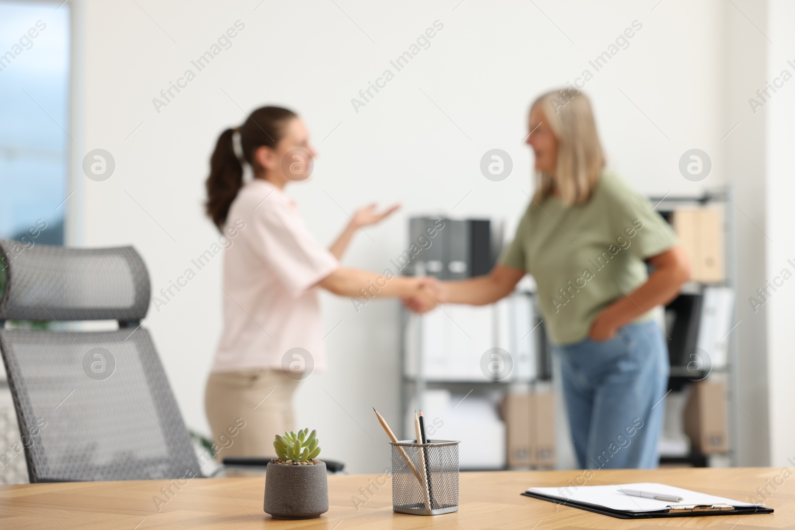 Photo of Women shaking hands in office, focus on wooden table with clipboard and stationery
