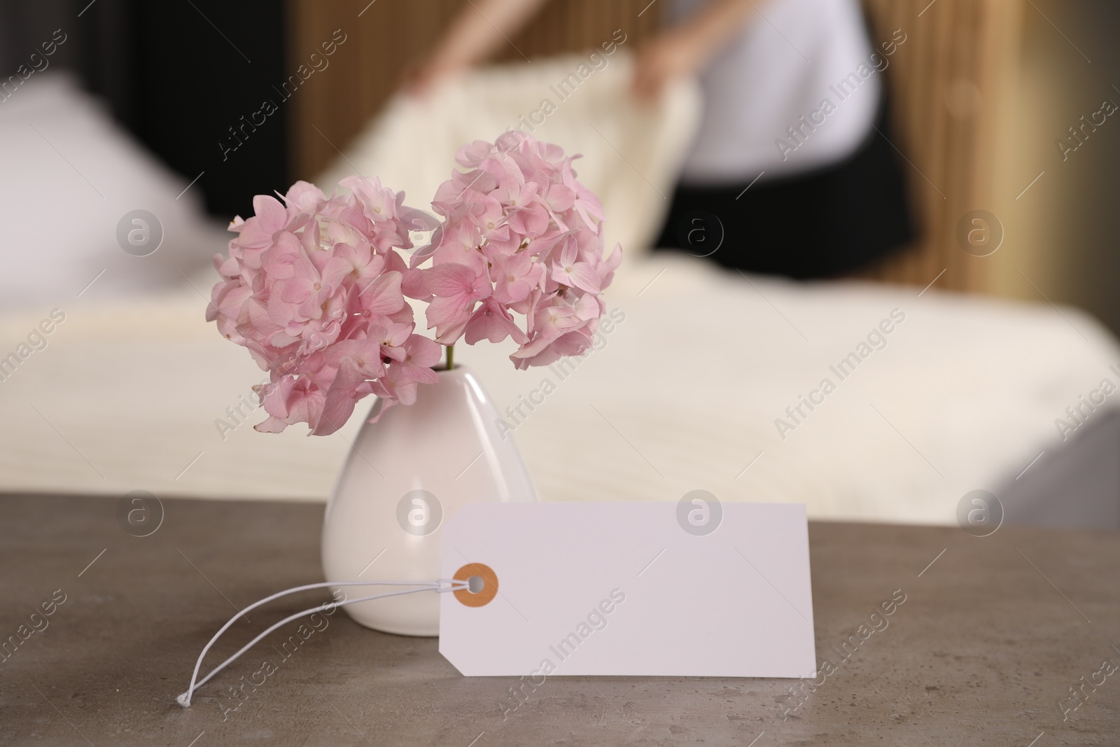 Photo of Welcome. Chambermaid making bed in hotel room, focus on blank card and hydrangea flowers