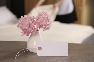 Photo of Welcome. Chambermaid making bed in hotel room, focus on blank card and hydrangea flowers