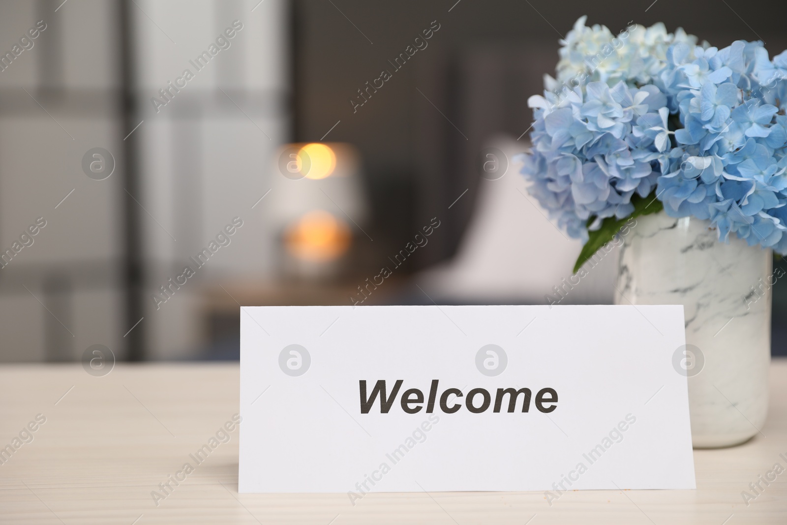 Photo of Card with word Welcome and hydrangea flowers on wooden table, closeup