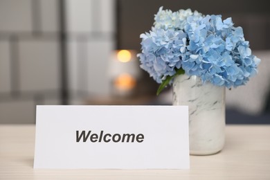 Photo of Card with word Welcome and hydrangea flowers on wooden table, closeup
