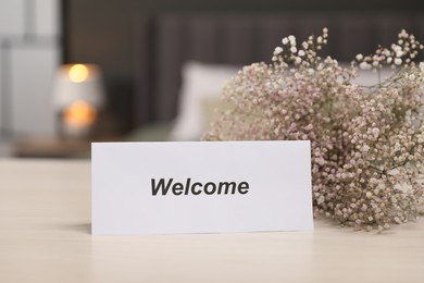 Photo of Card with word Welcome and gypsophila flowers on wooden table indoors, closeup