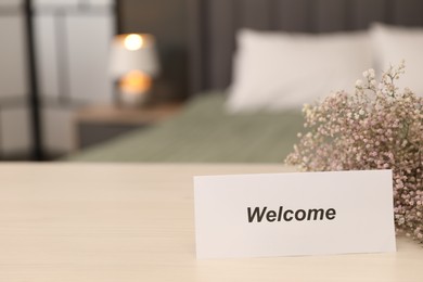 Photo of Card with word Welcome and gypsophila flowers on wooden table indoors, closeup. Space for text