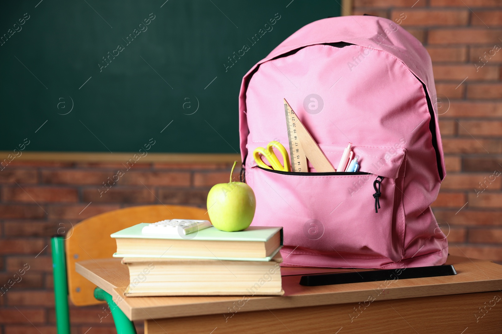 Photo of Pink backpack with different stationery, books and apple on desk in classroom, closeup
