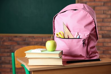 Photo of Pink backpack with different stationery, books and apple on desk in classroom, closeup