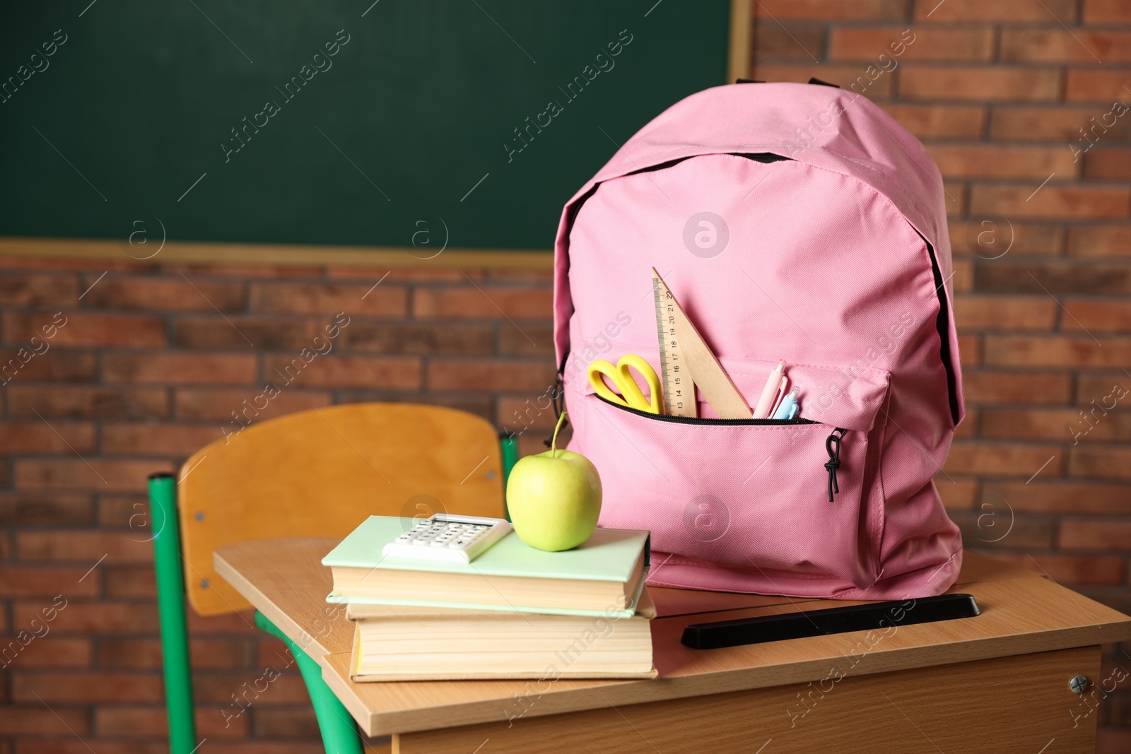 Photo of Pink backpack with different stationery, books and apple on desk in classroom, closeup