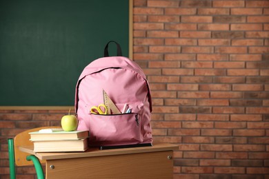 Photo of Pink backpack with different stationery, books and apple on desk in classroom. Space for text