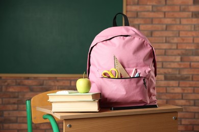 Photo of Pink backpack with different stationery, books and apple on desk in classroom