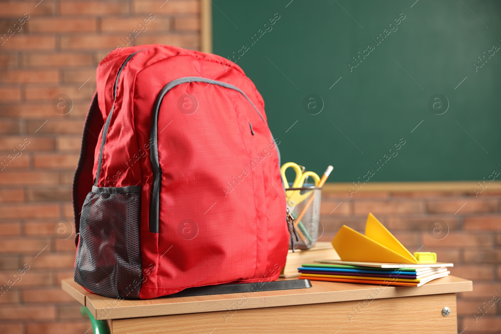 Photo of Red backpack, different stationery and paper plane on desk in classroom