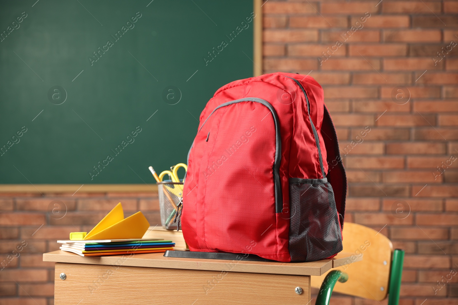 Photo of Red backpack, different stationery and paper plane on desk in classroom
