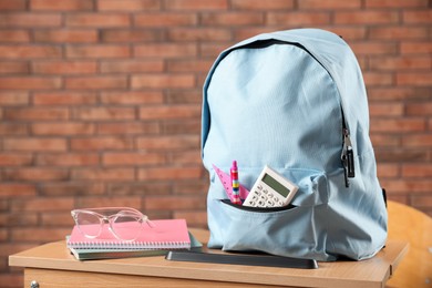 Photo of Light blue backpack with different stationery and glasses on desk in classroom