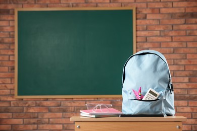 Photo of Light blue backpack with different stationery and glasses on desk in classroom. Space for text