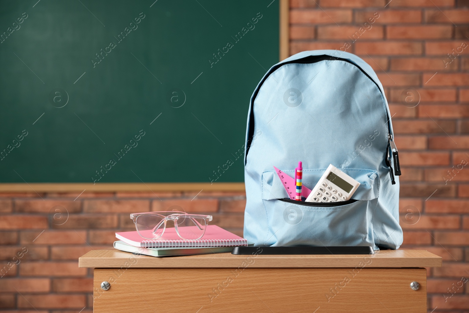 Photo of Light blue backpack with different stationery and glasses on desk in classroom