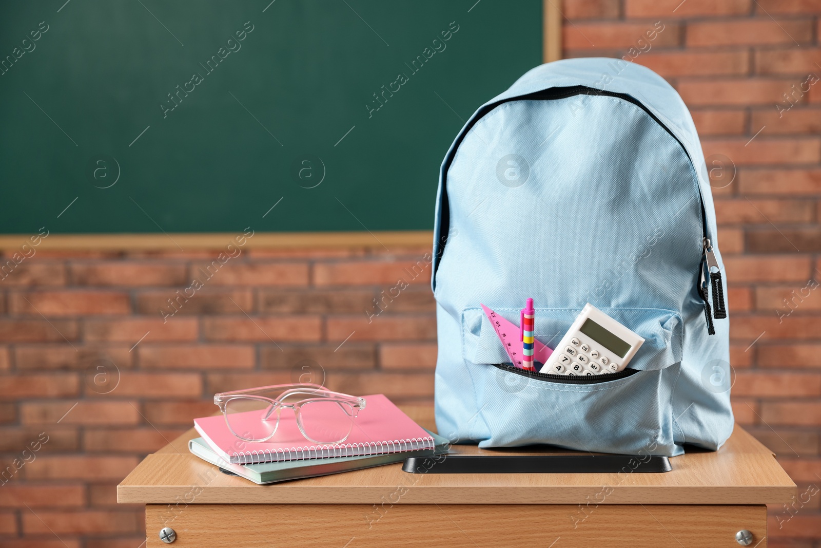 Photo of Light blue backpack with different stationery and glasses on desk in classroom