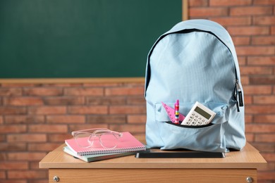 Photo of Light blue backpack with different stationery and glasses on desk in classroom