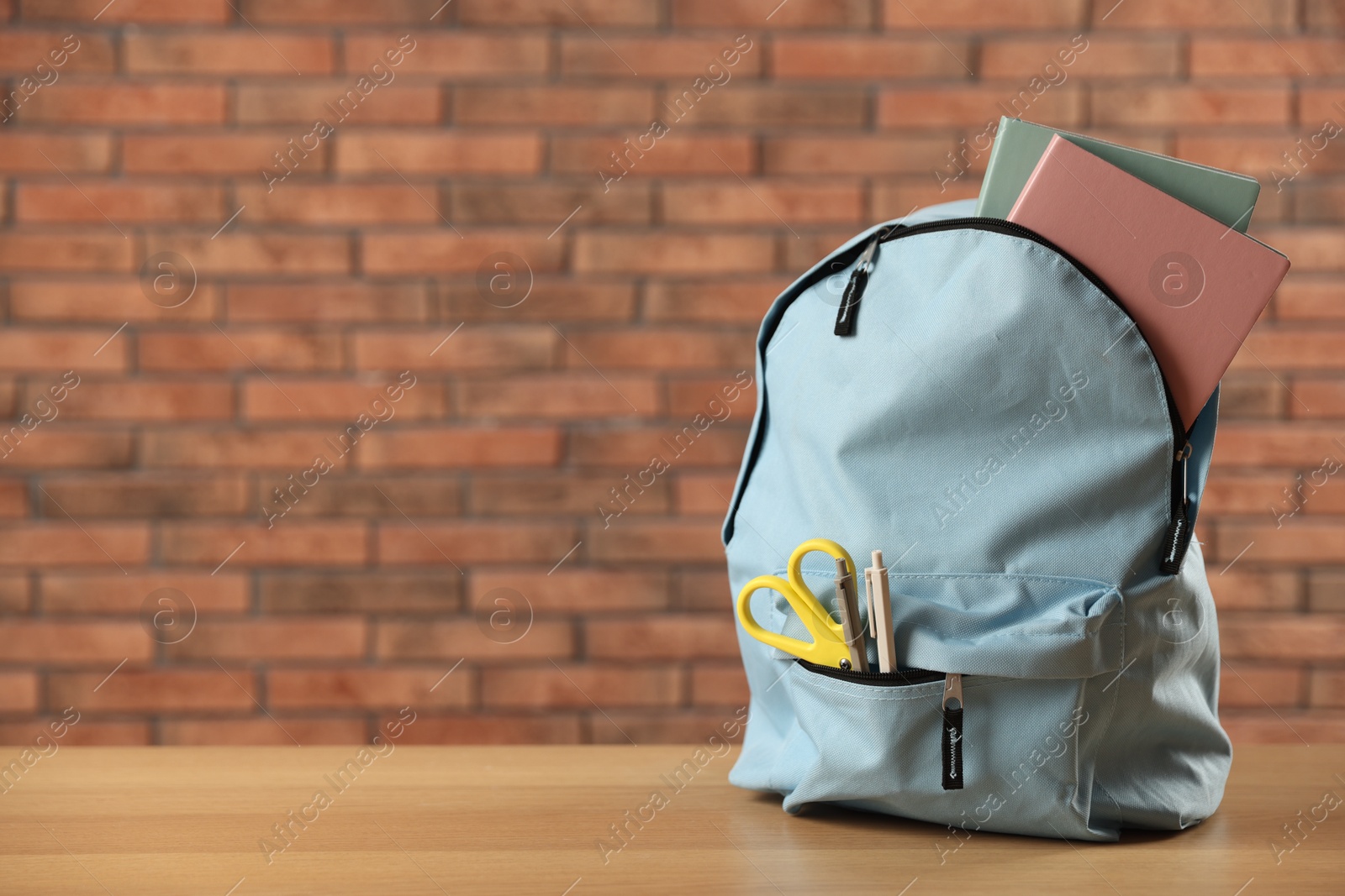 Photo of Light blue backpack with different stationery on wooden table against red brick wall. Space for text
