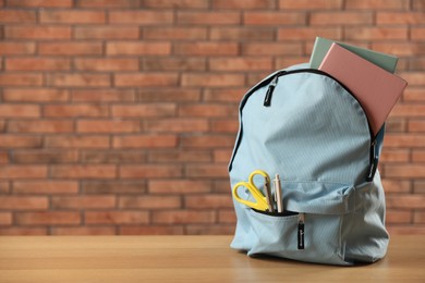 Photo of Light blue backpack with different stationery on wooden table against red brick wall. Space for text