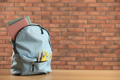 Photo of Light blue backpack with different stationery on wooden table against red brick wall. Space for text
