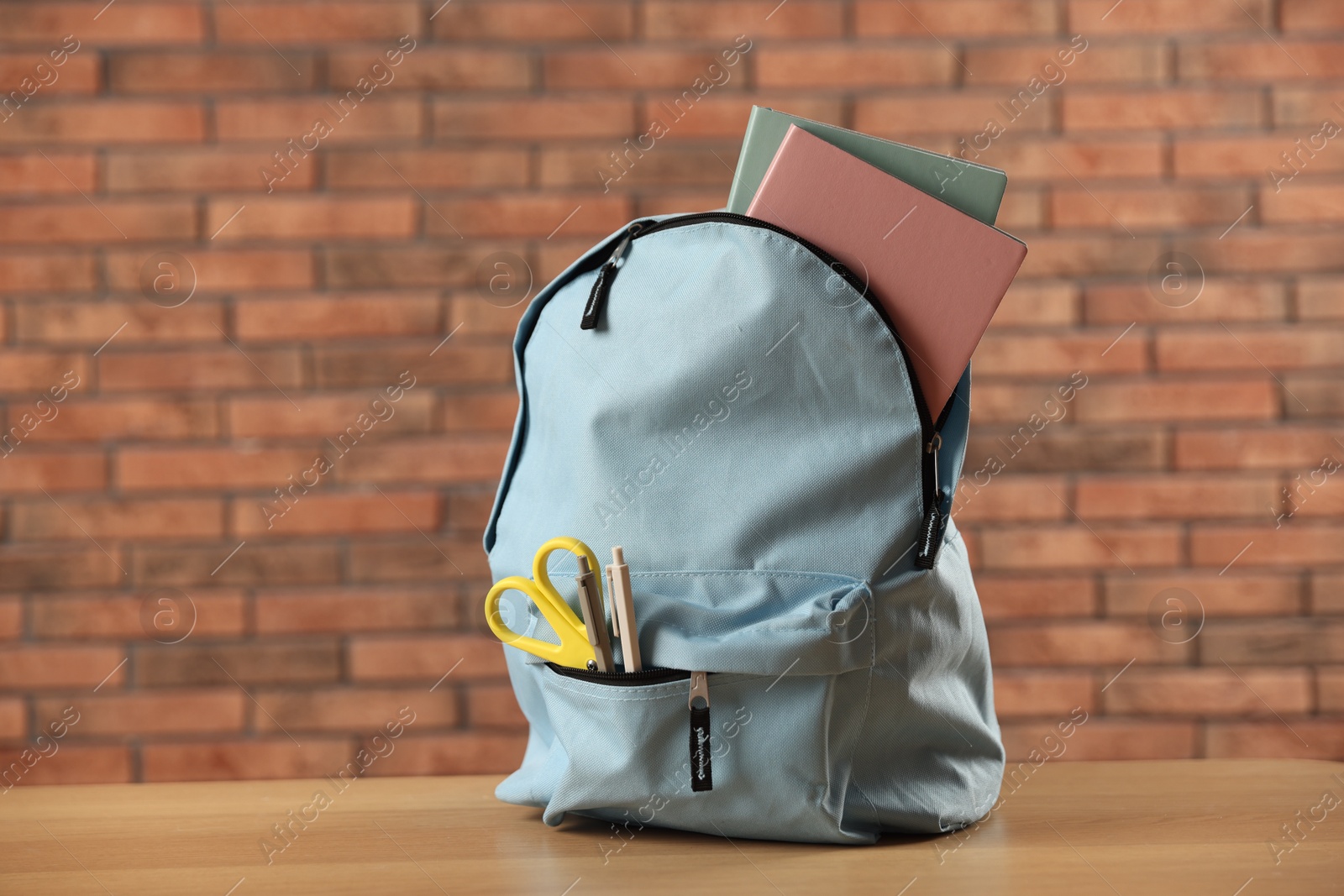 Photo of Light blue backpack with different stationery on wooden table against red brick wall
