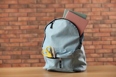 Photo of Light blue backpack with different stationery on wooden table against red brick wall