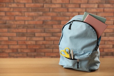 Photo of Light blue backpack with different stationery on wooden table against red brick wall. Space for text