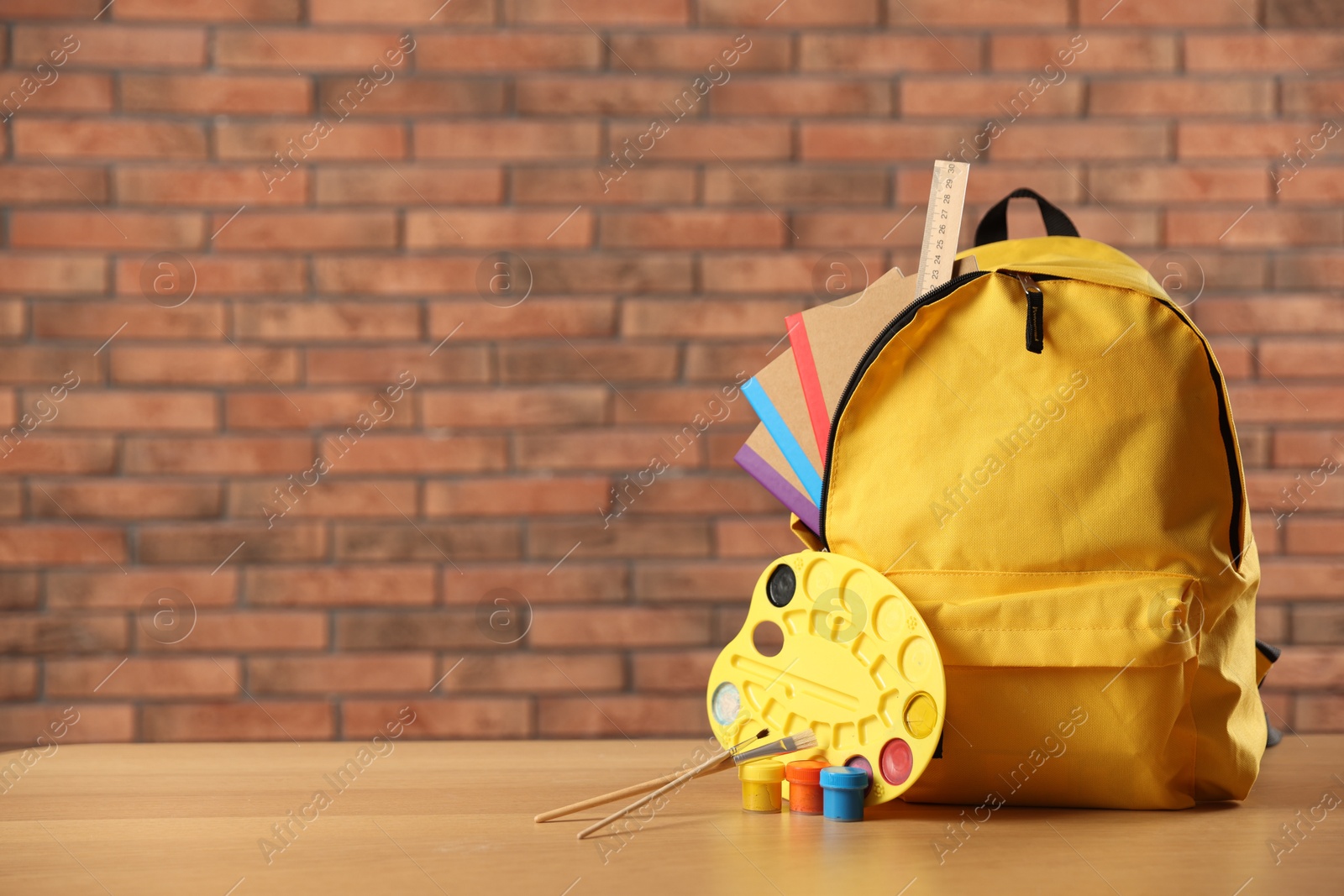 Photo of Yellow backpack with notebooks and art supplies on desk in classroom. Space for text