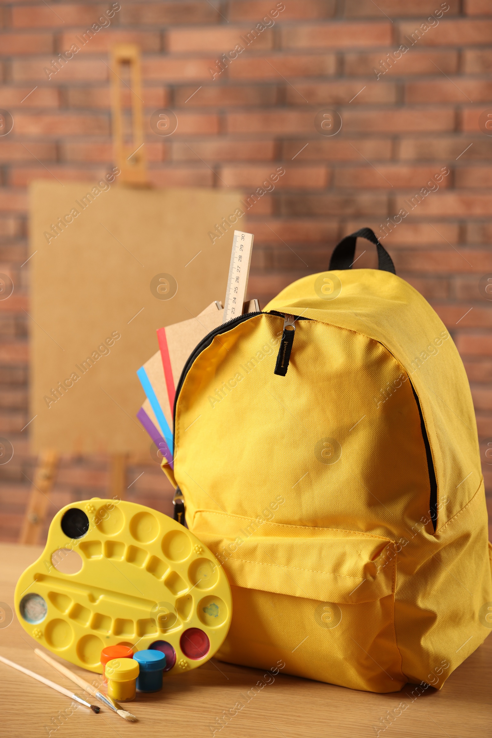 Photo of Yellow backpack with notebooks and art supplies on desk in classroom, closeup