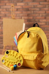 Photo of Yellow backpack with notebooks and art supplies on desk in classroom, closeup