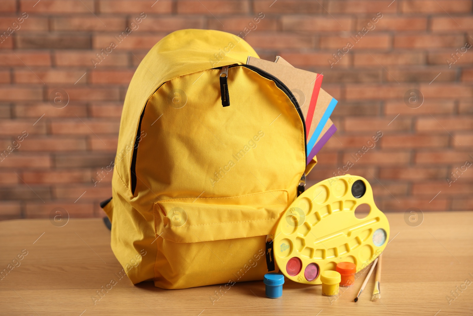 Photo of Yellow backpack with notebooks and art supplies on desk in classroom, closeup
