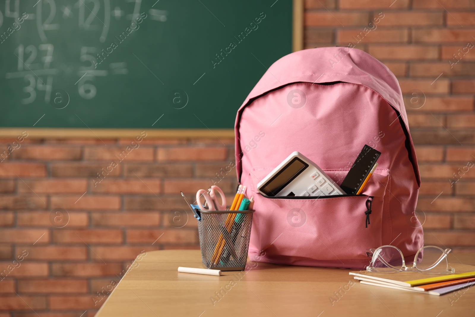 Pink backpack with different stationery on desk in classroom Photo of Pink backpack with different stationery on desk in classroom