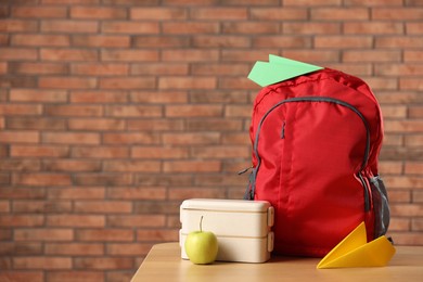 Photo of Backpack, paper planes, lunchbox and apple on wooden table against red brick wall. Space for text